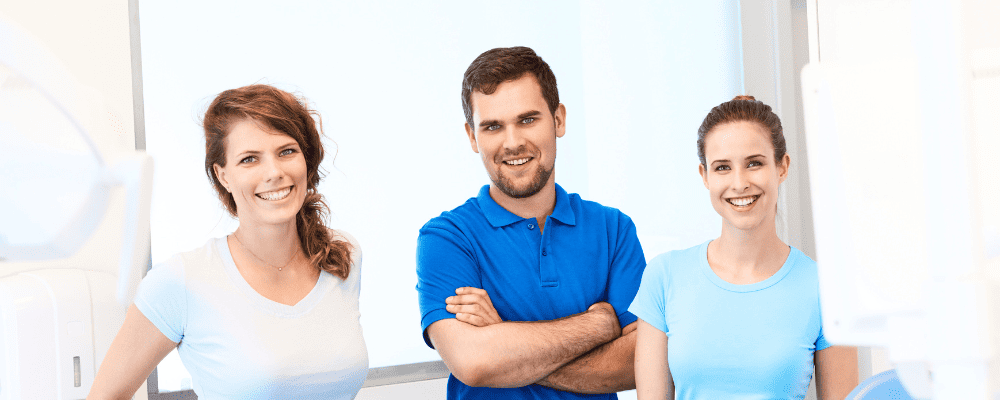 Zahnarzt zeigt sich mit stolzer Brust und seinen zwei ZFAs, die sich ungeplant bei ihm beworben haben. Team steht im Behandlungszimmer