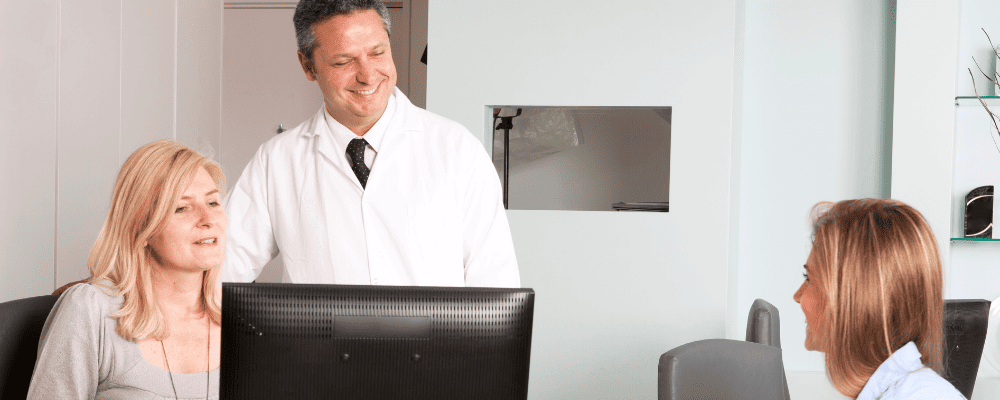 Älterer Inhaber einer Zahnarztpraxis in Kittel und mit Krawatte erklärt gemeinsam mit der Sekretärin einer jungen Mitarbeiterin, was für Social Media geplant ist